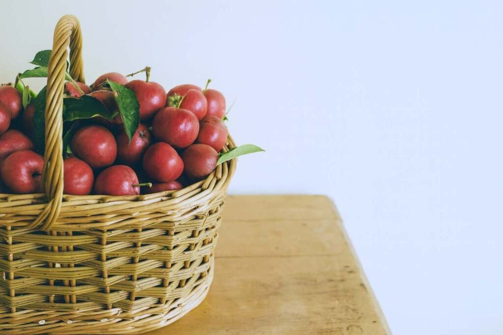 Woven wicker basket filled with red apples and leaves on a wooden table. Natural home harvest décor.