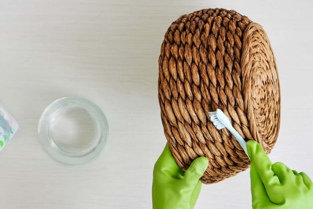 Gloved hands cleaning the bottom of a coiled seagrass basket with a toothbrush and water bowl.