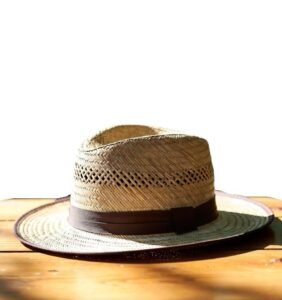 Classic tan straw fedora hat with a brown ribbon band resting on a sunlit wooden table.