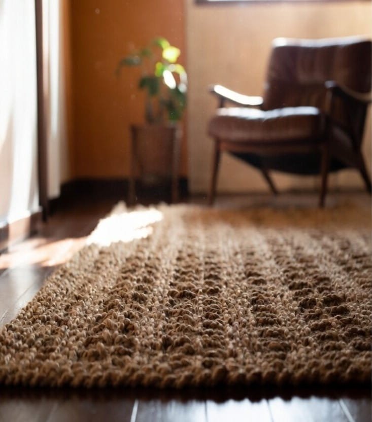 Close-up of a thick, textured jute rug on dark hardwood floor with a blurred leather chair in the background.
