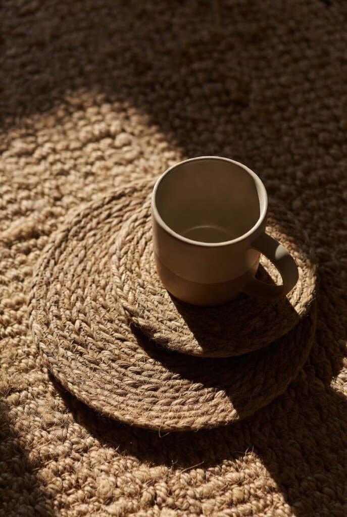 Stack of round braided jute coasters with a ceramic coffee mug on a textured natural fiber rug.