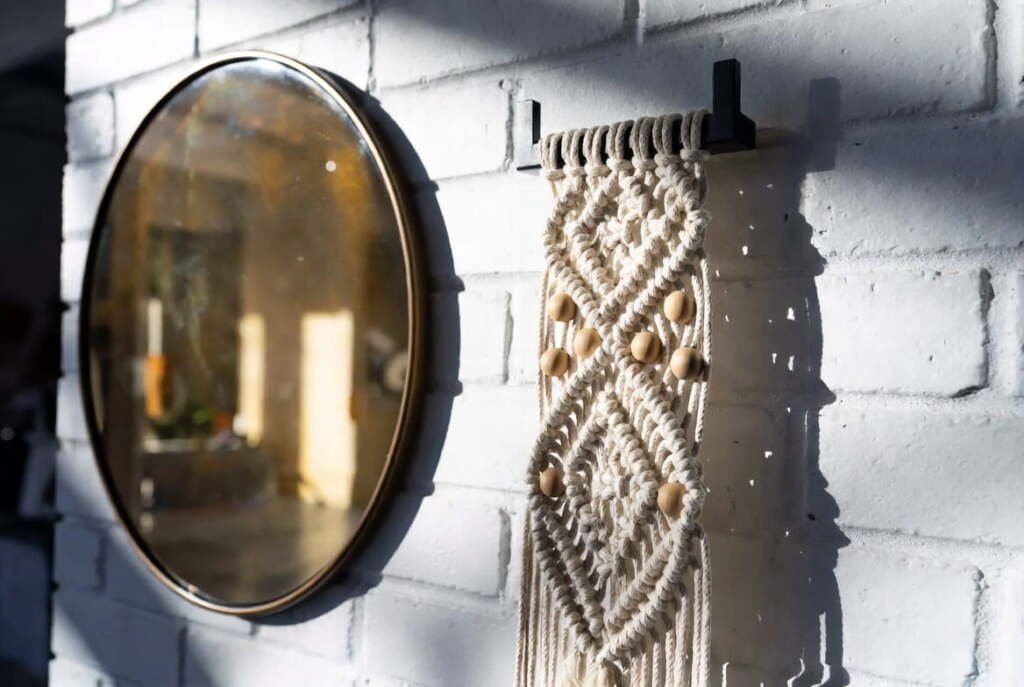 Close-up of modern macramé wall hangings with wooden beads on a white brick wall next to a round gold mirror.
