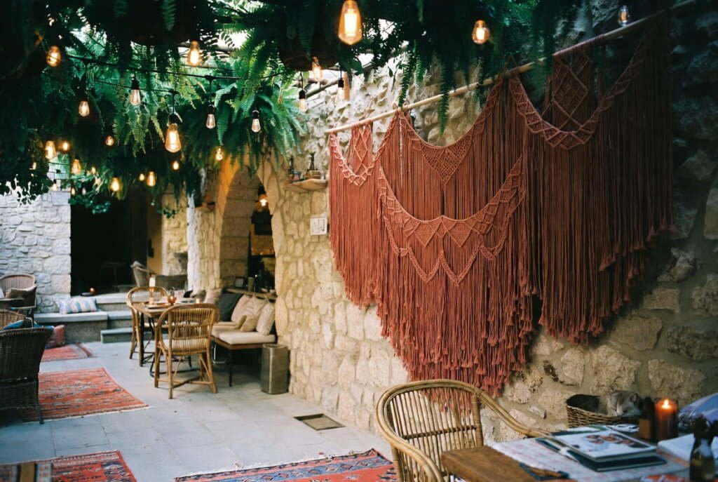 Large rust-colored macramé wall hangings on a stone patio wall under hanging ferns and warm string lights.