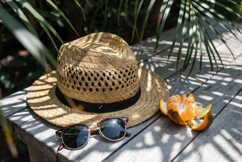 Close-up of a woven straw hat for summer travel, sunglasses, and orange on a wooden table with palm leaf shadows.