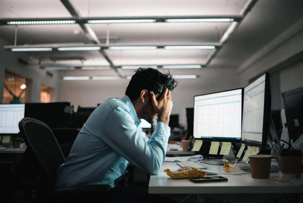 Stressed employee with head in hands at a cluttered office desk, representing workplace burnout and mental exhaustion.
