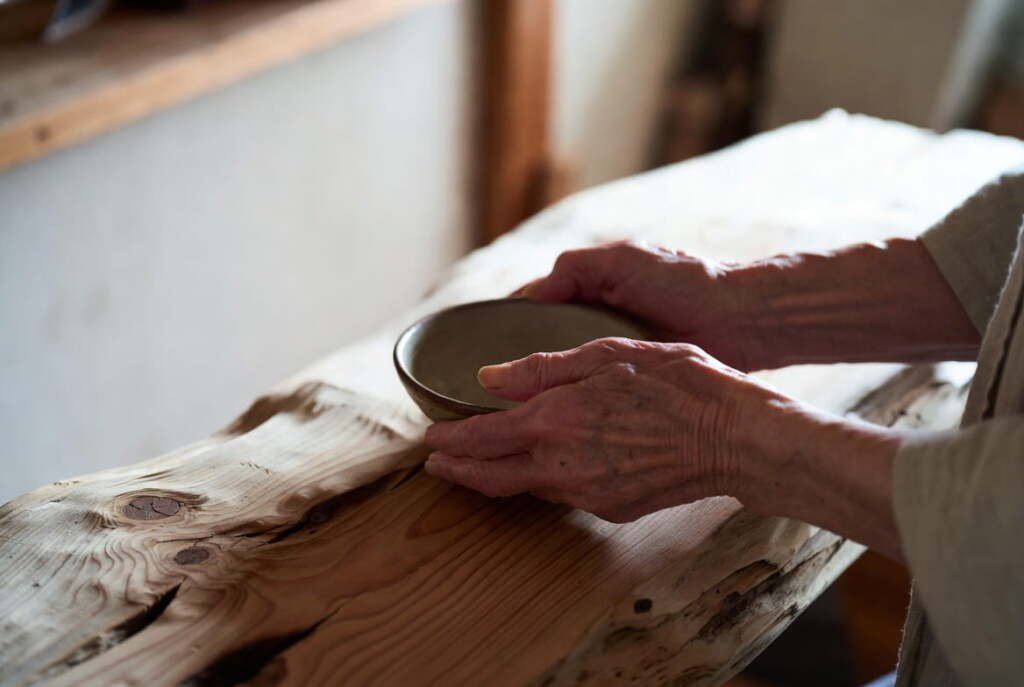 Elderly hands resting a ceramic bowl on a raw wood shelf — wabi-sabi and natural materials in a Japanese minimalist home