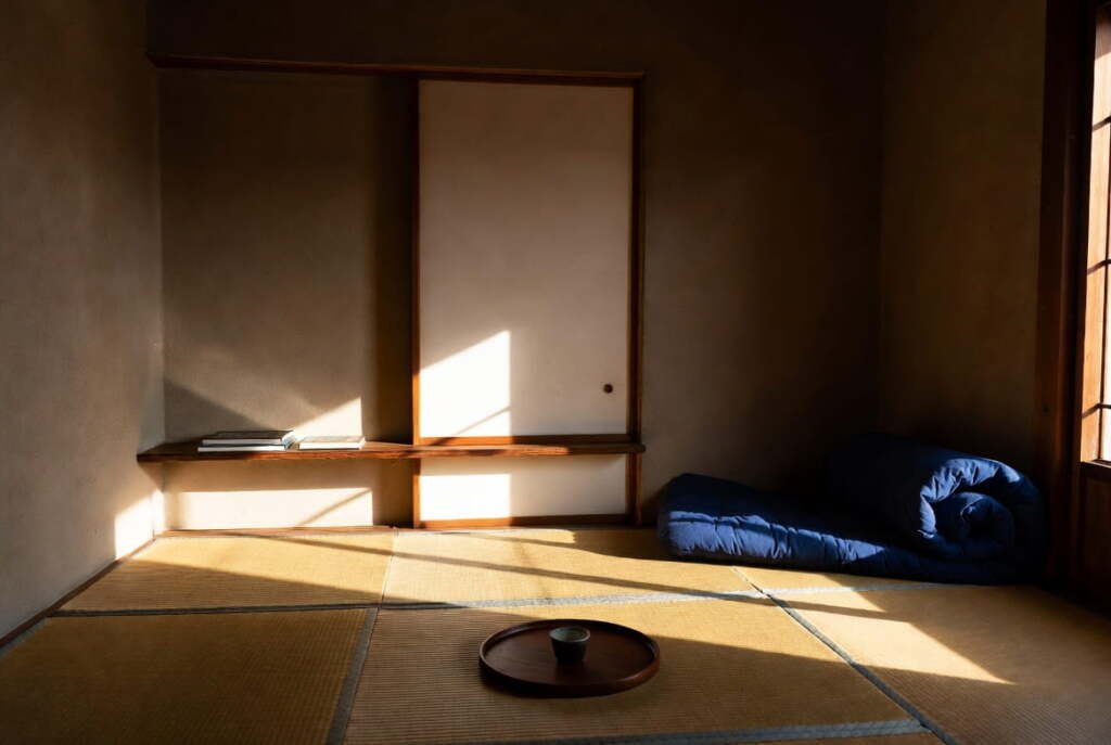 Japanese minimalist home tatami bedroom with soft morning light, folded blue futon, and a single tea tray on the floor