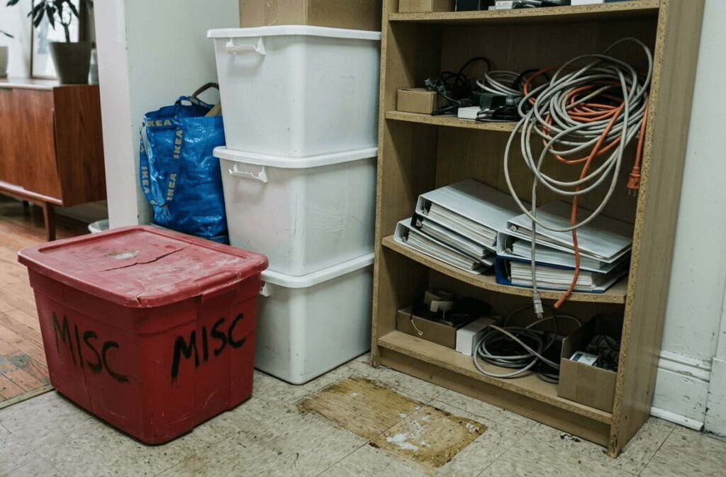 Mismatched plastic storage bins labeled MISC stacked next to a sagging particleboard shelf in an apartment corner