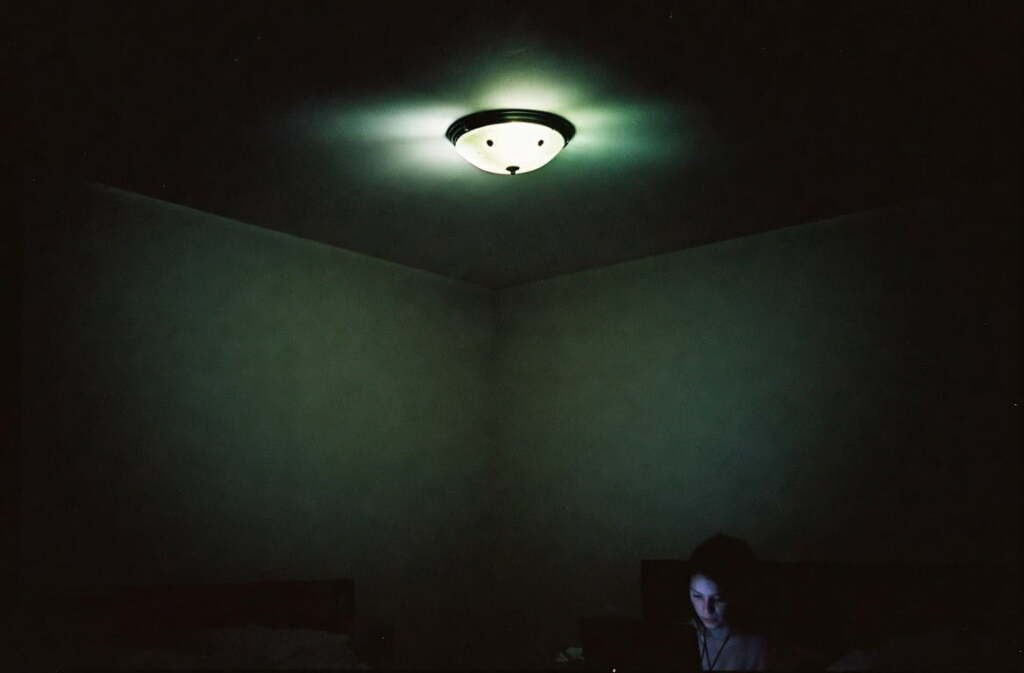 Woman working at desk in a dark room lit only by one harsh overhead ceiling light with deep shadows in every corner